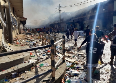 People gather as smoke rises from a burnt out buildings in Honiara's Chinatown on November 26, 2021 after two days of rioting which saw thousands ignore a government lockdown order, torching several buildings around the Chinatown district including commercial properties and a bank branch.