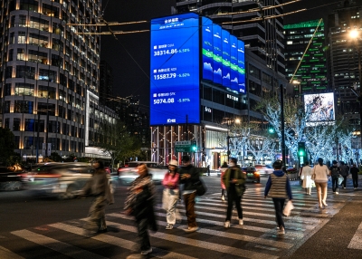 People walk next to a screen showing the global index, including Dow Jones, NASDAQ and the S&P global ratings, in the Jing'an district in Shanghai on April 7, 2025.